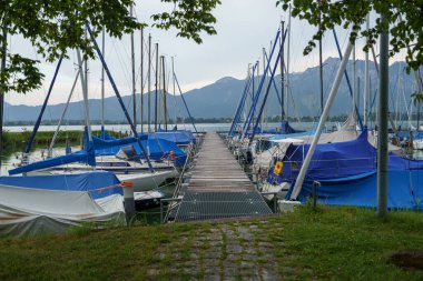 Mavi kapaklı yelkenliler marinaya yanaştı. Sakin su akşam gökyüzünü yansıtır. Arka planda dağlar belirir ve alacakaranlıkta huzurlu bir atmosfer yaratır..