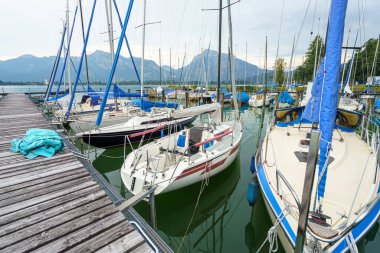 Birçok yelkenli, arka planda dağlar olan sakin bir gölün limanına yanaştı. Kara bulutlar olası yağmuru işaret ediyor, barışçıl ama dramatik bir sahne yaratıyor..