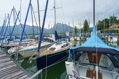 Yat limanında çok sayıda yelkenli var. Suyun üzerinde yavaşça sallanıyorlar. Arkaplanda güneş batarken kasvetli bir gökyüzünün altında görkemli dağlar var..