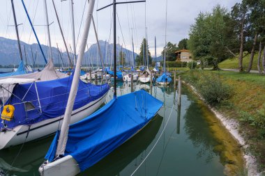 Mavi kapaklı birkaç yelkenli tekne dağlarla çevrili sakin bir marinaya yanaştı. Atmosfer sakin ve gökyüzü bulutlu. Sahnenin doğal güzelliğini arttırıyor..