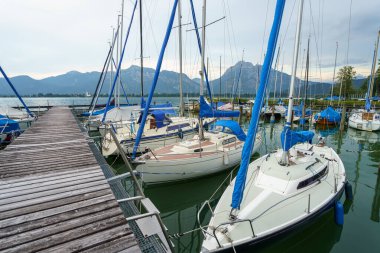 Birkaç yelkenli marinaya ahşap geçitli demirlemiş. Sahnede akşam erken saatlerde bulutlu bir gökyüzünün altında arka planda dağlar yer alıyor..