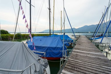 Renkli kapaklı yelkenliler huzurlu bir marinaya demirlediler. Etrafındaki dağlar ve bulutlu gökyüzü sakin bir atmosfer yaratıyor. Su kenarında rahatlatıcı bir gün için mükemmel..