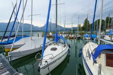 Renkli yelkenliler dağlarla çevrili huzurlu bir marinaya demirlediler. Su bulutlu gökyüzünü yansıtır ve öğleden sonra erken saatlerde sakin bir atmosfer yaratır..