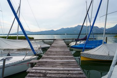 Sabahın erken saatlerinde huzurlu bir marinada tahta iskeleye yanaşmış birkaç yelkenli görülüyor. Arkaplanda yemyeşil dağlar yükselir bulutlu bir gökyüzünün altında.