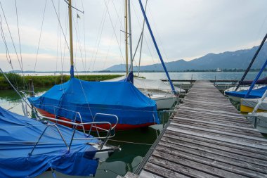 Mavi kapaklı yelkenliler, su kıyısındaki ahşap bir rıhtıma demirlediler. Ufuk boyunca uzanan dağlar bulutlu bir gökyüzünün altında. Huzurlu ve huzurlu bir sahne..