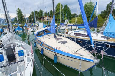 Birden fazla yelkenli, bulutlu bir gökyüzünün altında arka planda yeşil ağaçlarla dolu canlı bir marinada demirli. Sahne huzurlu bir dinlenme ve kayık atmosferi yakalıyor..