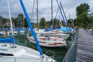Yelkenli koleksiyonu bir marinaya demirlemiş, yelkenleri mavi örtülerle kaplanmış. Arka planda bulutlu gökyüzünün altında ağaçların oluşturduğu sahne huzur verici..