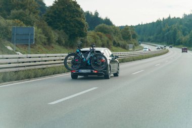 Bir araç arka taraftaki bisiklet parkına bağlı iki bisikletle düz bir otoyol boyunca sürer. Manzara ağaçlar ve açık bir gökyüzü içerir, açık hava macera atmosferini arttırır..