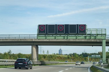 Araçlar yoğun bir otoyolda seyahat ederken dijital hız limiti tabelaları saatte 100 km 'yi gösteriyor. Sahne, açık bir gökyüzünü ve arka planda güneşli bir öğleden sonranın tipik yeşilliğini gösteriyor..