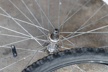 Bir bisiklet çarkı beton zeminde durur ve merkezinin ve tellerinin karmaşık tasarımını ortaya çıkarır. Bu detaylı görüntü, metal ve lastik bileşenleri bir garaj ayarında yakalıyor..
