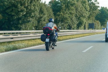 Bir motosikletçi otoban boyunca kask ve turuncu giysiler giyerek gezer. Bisiklet yan çantalarla donatılmıştır ve ağaçlar güneşli bir günde yol kenarına dizilmiştir..
