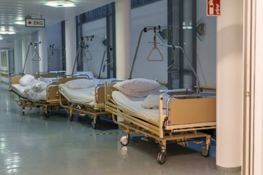 Sessiz bir hastane koridoru duvarın karşısında birkaç boş yatak gösteriyor. Yataklar düzenli ve gelecekteki hastalar için hazır. Yumuşak ışık, sakin atmosfere katkı sağlar..