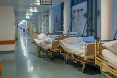 Sessiz bir hastane koridorunda, birkaç boş hasta yatağı duvar boyunca dizilmiştir. Tıbbi teçhizat görünür, steril ve düzenli bir alan gösteriyor. Işık sakin bir atmosfer yaratıyor..