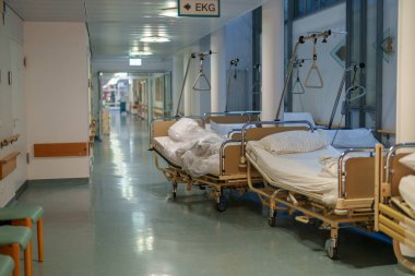 Olay yeri boş hasta yataklarıyla dolu sessiz bir hastane koridorunu gösteriyor. Yumuşak ışıklandırma, hasta ziyaretleri arasında sakin bir atmosfer yaratıyor..