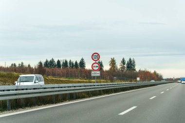 Araçlar otobanda maksimum hız limiti tabelasıyla seyahat ederler. Yol kenarına beyaz bir minibüs park edilmiş. Hava bulutlu ve etrafındaki ağaçlar sonbahar belirtileri gösteriyor..