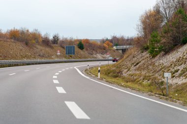 Gri bir asfalt otoban bulutlu bir günde Deutschland kırsalında kıvrılır. Kırmızı bir araba otobanda bir üst geçidin altında yol alır..