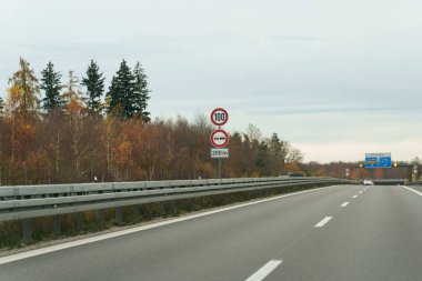Bulutlu bir gökyüzünün altında gri bir otoyol uzanıyor. Sonbahar ağaçları yol kenarına dizilmiş ve bariyerlerin yanında hız sınırı işareti var. Yalnız beyaz bir araba otobanda sürüyor..