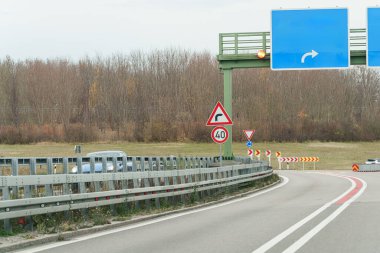 Gri bir günde, araçlar sonbahar ağaçlarının yanındaki bir otoyolda seyahat ederler. Yol kenarında birden fazla işaret var. Yol eğriliği hakkında yön ve uyarı ver..