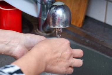 Bir insan mutfaktaki gümüş musluktan akan suyun altında ellerini yıkıyor. Elleri yıkanırken musluktan su dökülüyor..