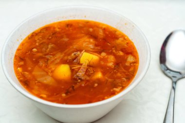 Yakın plan Ukrayna borşuyla dolu beyaz bir kase gösteriyor. Patates ve lahana parçaları görülebilir. Parlak bir kaşık kasenin yanında duruyor, yenmeye hazır olduğunu gösteriyor..