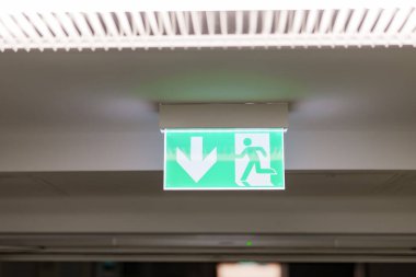 Parlayan yeşil bir işaret acil çıkışı gösteriyor. Sembol, kapıyı işaret eden bir okla kapıya doğru koşan birini gösteriyor. Kamu binasının tavanında asılı duruyor..