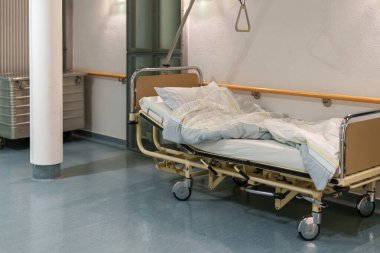 Boş bir hastane yatağı sessiz bir odada yastık ve battaniyeyle hazır bekliyor. Tıbbi teçhizat ve destek çubuğu görünüyor, sıradaki hastayı bekliyorlar..