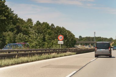 Arabalar bir Alman otoyolunda gidiyor. Bir hız sınırı işareti gösterilir. Ağaçlar yol kenarında bulutlu, aydınlık bir günde, kırsal kesime bir bakış sunuyor..