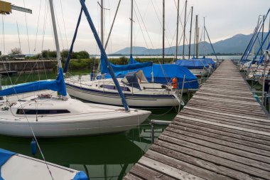 Starnberger See 'deki ahşap rıhtımda birkaç yelkenli gemi demirlemiş. Teknelerin mavi yelkenleri ve örtüleri var. Dağlar arka planda bulutlu bir gökyüzünün altında görülebilir..