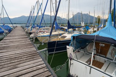 Bir grup yelkenli Chiemsee Gölü 'ndeki eski ahşap iskeleye demirlemiş. Arka planda dağlar yükselir, Prien 'de sakin ve resim gibi bir manzara yaratır..