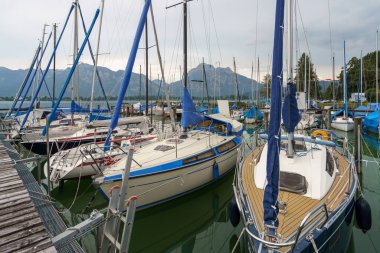 Yelkenliler bir limanın sakin sularında dinlenir. Direkler Tegernsee 'de bulutlu bir gökyüzünün altında uzak dağların arka planına karşı yükselir..