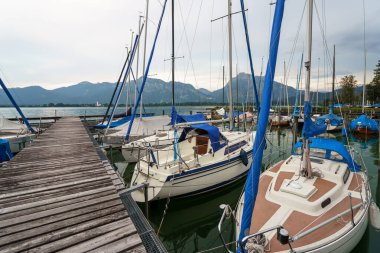 Yelkenliler Tegernsee 'ye yanaştı. Dağlar bulutlu bir gökyüzünün altında yükselir. Tahta bir iskele demirli gemilere gidiyor. Su sakin..