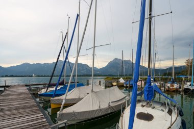 Yelkenliler bulutlu bir günde Chiemsee Gölü 'ndeki bir rıhtıma demirlerler. Arka planda dağlar yükselir. Bazı tekneler korunuyor, onları elementlerden koruyor..
