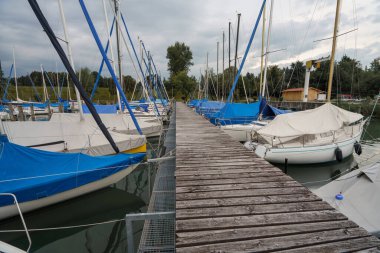 Starnberger See 'deki rıhtımlı yelkenlilerin arasında uzun bir ahşap iskele uzanır. Yelkenler açık, örtüler teknelerde, gölde bir sonraki yolculuklarını bekliyorlar..
