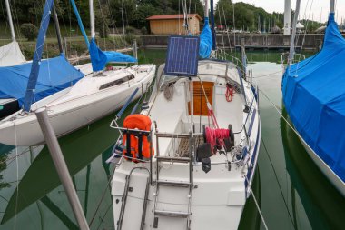 Bir marinada üç yelkenli yan yana yanaşmış. Tekneler mavi detaylarla bembeyaz ve birinde güneş paneli var. Su yeşil, etrafındaki ağaçları ve gökyüzünü yansıtıyor..
