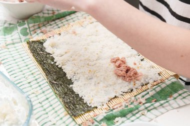 Bir kişi evde suşi yapıyor. Pişmiş pirinç bir nori çarşafına yayılır. Pirincin üstüne konserve ton balığı ekleniyor..