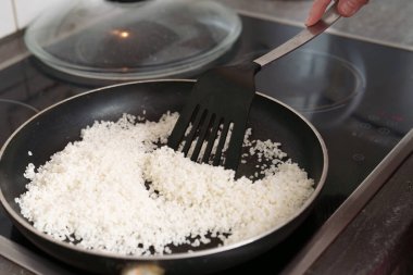 Birisi siyah bir tavada beyaz pirinç karıştırıyor. Mutfak sobasında siyah bir spatula. Pirinç, yemek pişirmek ve istenen dokuya ulaşmak için etrafa saçılıyor..