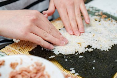 İki el, suşi rulosu yapmaya hazırlanmak için bambu suşi hasırında kurutulmuş yosunların üzerinde pişmiş beyaz pirinci dikkatlice yayıyor..