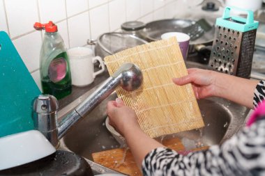Bir insan mutfak lavabosunda bambu suşi paspası yıkıyor. Bulaşıklar ve mutfak aletleri lavabonun etrafında. Sahne ışıl ışıl aydınlandı, ev işlerini gösteriyor..