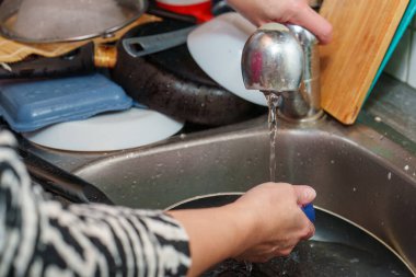 Elleri paslanmaz çelik bir lavaboda tavayı fırçalamakla meşgul. Bulaşıklar ve tencereler lavaboya taşar. Musluk suyla çalışır. Mutfağı temizlemek için çok çalışıyor..