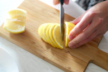 Yakın plan, ahşap bir kesme tahtasıyla parlak sarı bir limonu dilimleyen elleri gösteriyor. Dilimler düzgünce düzenlenir, ferahlatıcı bir içecek ya da aşçı kreasyonu için hazırdır..
