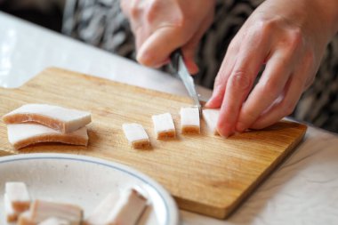 Yakından bakıldığında, parlak ışıklandırılmış bir mutfakta yemek hazırlama sırasında ahşap bir kesme tahtasının üzerinde domuz yağını küçük küplere ayıran eller görülüyor..