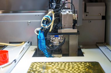 A machine operates in a factory, focusing on a specific task. Various tubes and wires connect to the device. The scene shows the complexity of industrial production.