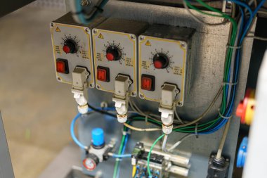 Metal bir panele üç tane kontrol anahtarı monte edilmiş. Her anahtarın göstergeleri ve bağlantıları var. Kablolar endüstriyel makine ortamında farklı bileşenlere yol açar..