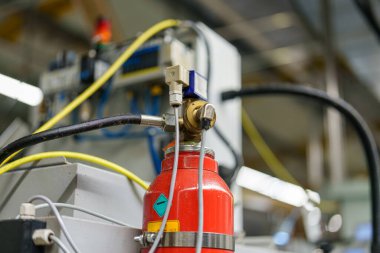 Bir fabrikada yangın söndürme sistemi kuruldu. Ekipman kırmızı silindir ve kablolu kontrol panelini içeriyor. İşçiler var ama görünür değiller..