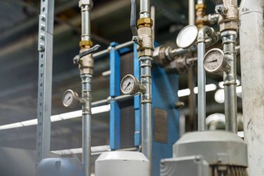 İşçiler borular ve göstergelerle dolu bir fabrikada çeşitli aletler kullanıyorlar. Ölçüler ve vanalar gibi ekipmanlar görünür, basınç seviyelerini ve çalışma koşullarını gösterir.