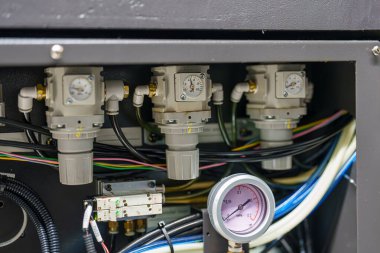 Ekipman kontrolleri üç basınç düzenleyicisi ve bir gösterge gösteriyor. Kurulumda çeşitli kablolar ve montajlar yer alıyor. Bu muhtemelen bir fabrikadaki ya da atölyedeki bir makinenin parçası..