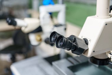 Bir mikroskop laboratuvar masasına yakın aletlerle yerleştirildi. Optik lens görünür ve arka plan daha fazla laboratuvar ekipmanı ve çalışma alanını gösteriyor..