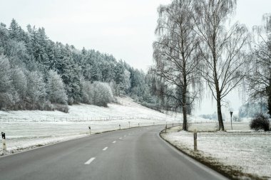 Her iki tarafında ağaçlar olan karlı bir arazide dolambaçlı bir yol var. Gökyüzü bulutlu ve yer beyaz karla kaplı. Kırsal bir ortam sessiz bir sahne yaratır..
