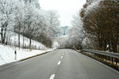 Her iki tarafında karla kaplı ağaçların olduğu bir kış sahnesinde bir yol uzanıyor. Sahne temiz bir yol ve kırsal bir bölgede gri gökyüzünün altında beyaz bir arazi gösteriyor..