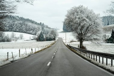 Arka planda ağaçlar ve tepeler olan beyaz karların içinden geçen uzun bir yol. Sahne bulutlu bir gökyüzünü ve sakin bir kırsal alanı gösteriyor..